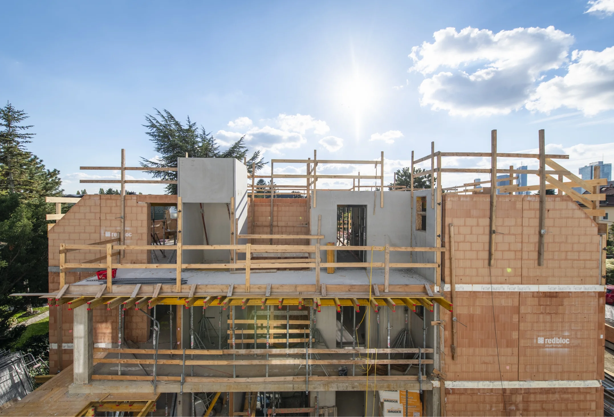 Frontale Baustellenansicht des obersten Stockwerks eines Rohbaus im hellen Sonnenlicht, die Wände aus rotem Ziegelmauerwerk, Betonelemente und hölzerne Absturzsicherungen zeigt.