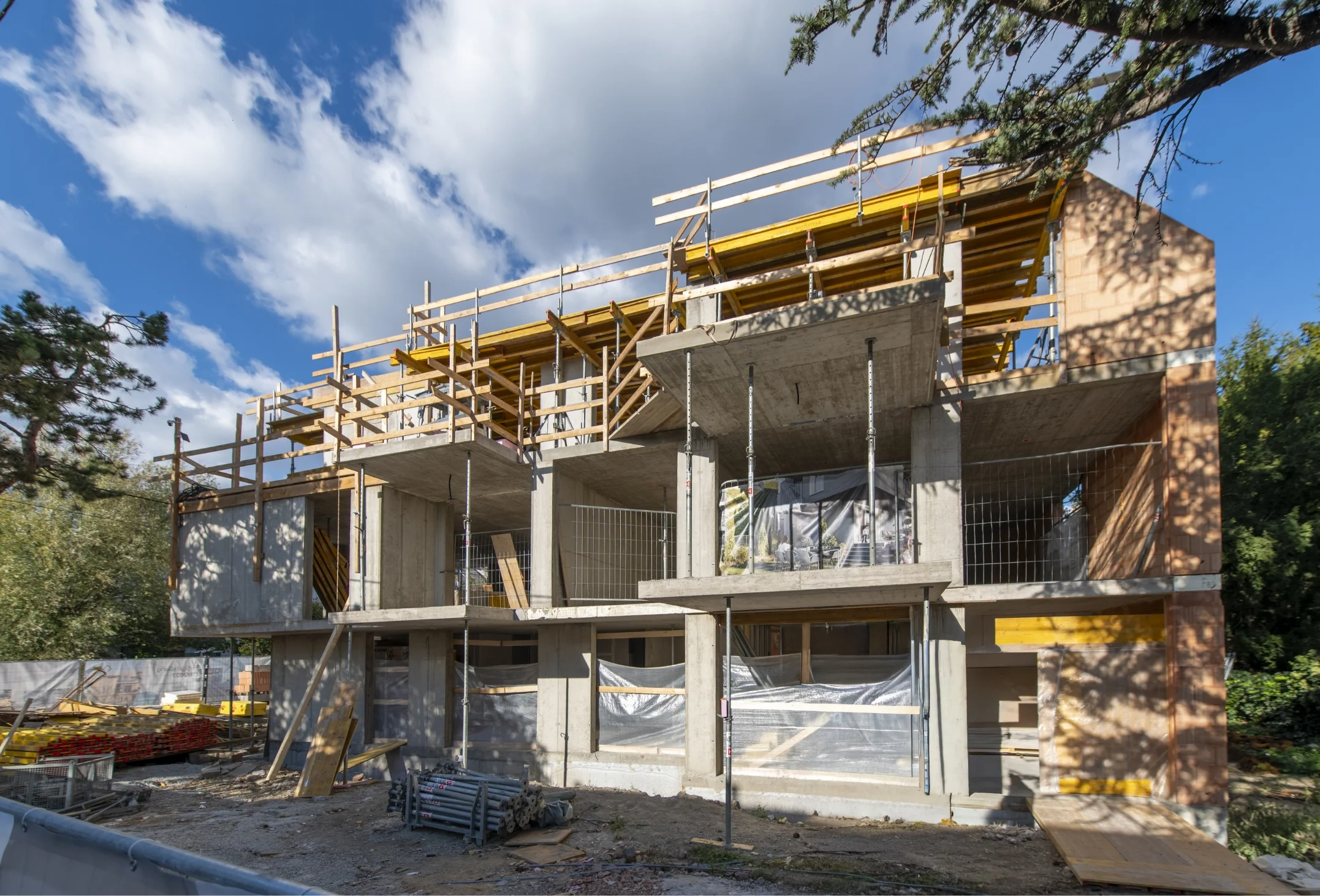 Außenansicht eines mehrstöckigen Gebäudes im Rohbauzustand mit sichtbaren Betondecken, Metallstützen für die Balkone, Ziegelwänden und Holzverschalungen im Dachgeschoss.