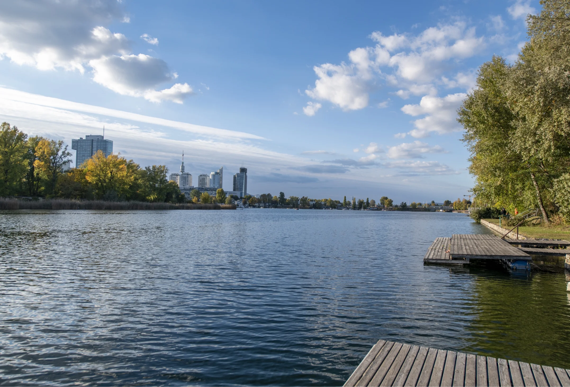 Landschaftsaufnahme eines ruhigen Gewässers mit Holzstegen im Vordergrund und einer städtischen Skyline im Hintergrund unter blauem Himmel.