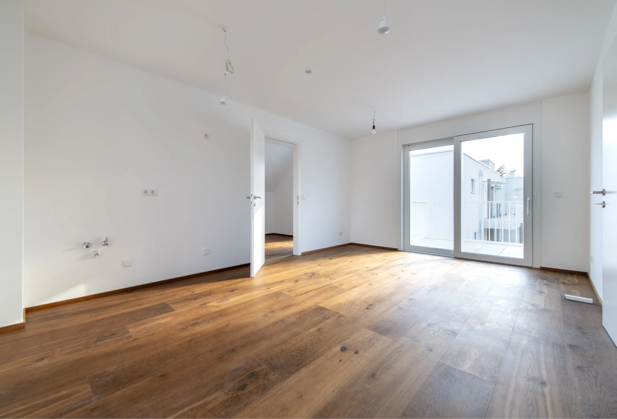 Heller, leerer Innenraum mit Holzboden, weißen Wänden, Anschlüssen für eine Küche und einer großen Glastür, die auf einen Balkon führt.