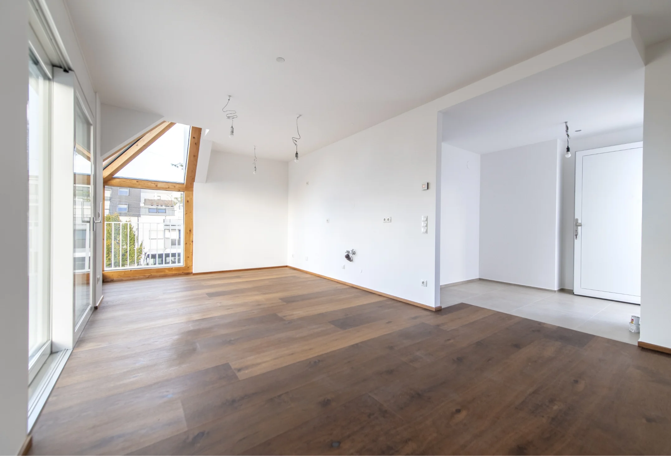 eerer Wohnraum mit Holzboden, weißen Wänden und einem markanten, teilweise schrägen Fenster mit Holzrahmen sowie einem Durchgang zu einem gefliesten Flur.