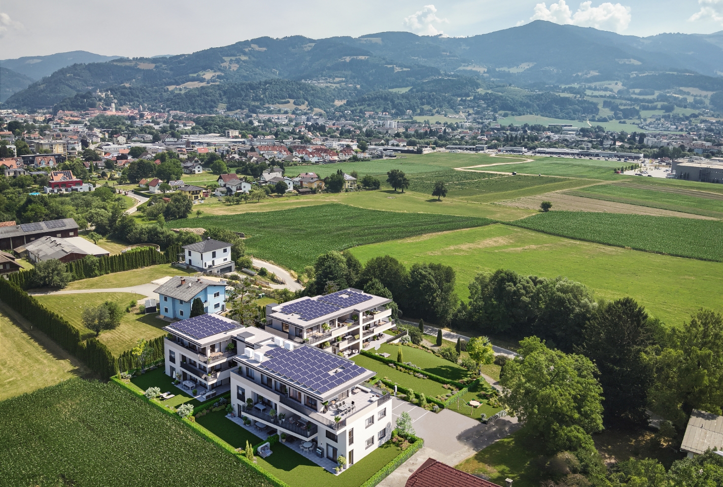 Drohnenaufnahme moderner Wohnanlagen mit PV-Dächern inmitten grüner Felder und Bergpanorama.