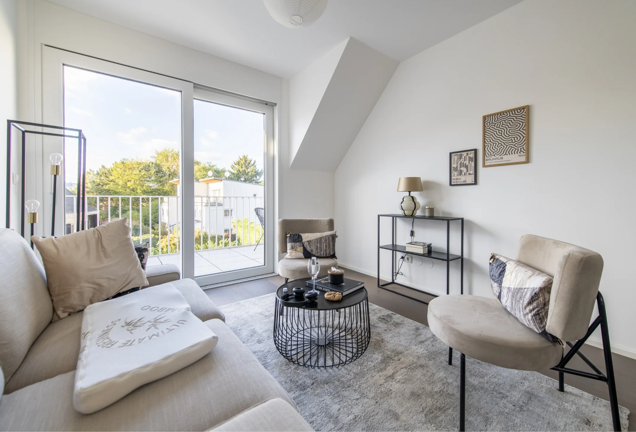 Ein modern eingerichtetes Wohnzimmer mit Dachschräge, das ein graues Sofa, Sessel, einen runden Couchtisch aus Metall und eine große Glastür zum Balkon zeigt.