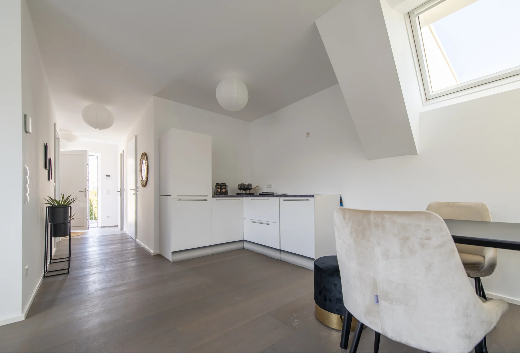 Ein offener Küchen- und Essbereich mit weißen, grifflosen Einbauschränken, dunklem Holzboden, einem Tisch mit hellen Stühlen und Blick in einen angrenzenden Flur.