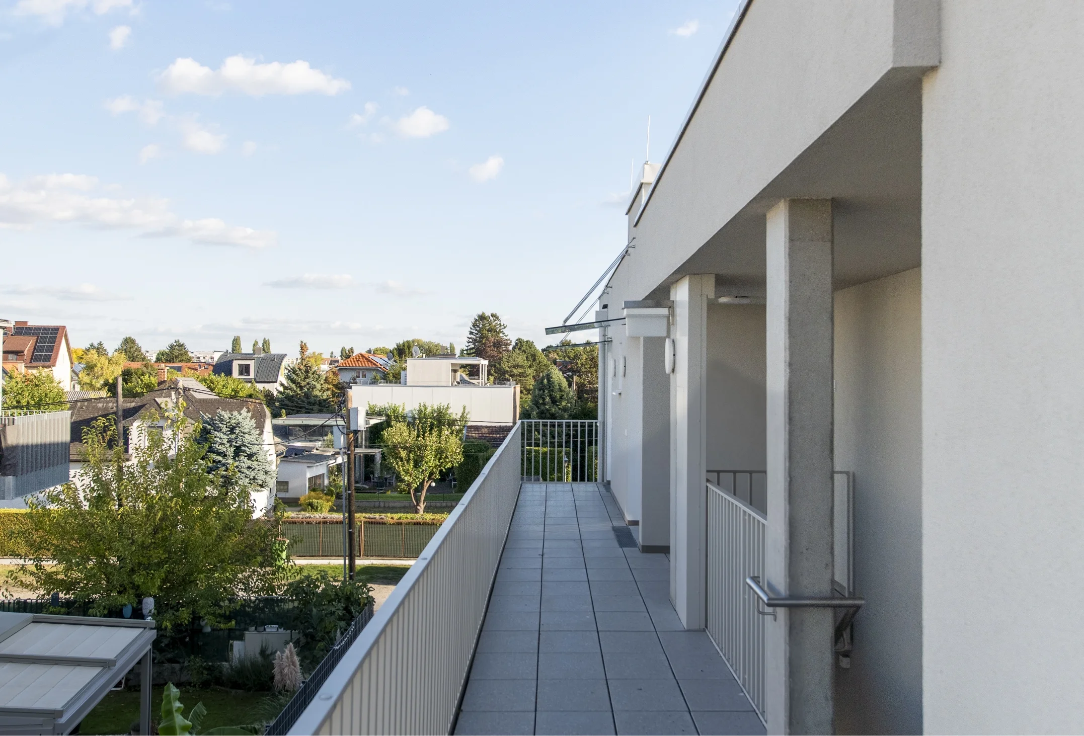 Ein langgestreckter, grau gefliester Balkon mit weißem Metallgeländer und Ausblick auf eine umliegende Wohnsiedlung mit Bäumen und Häusern.