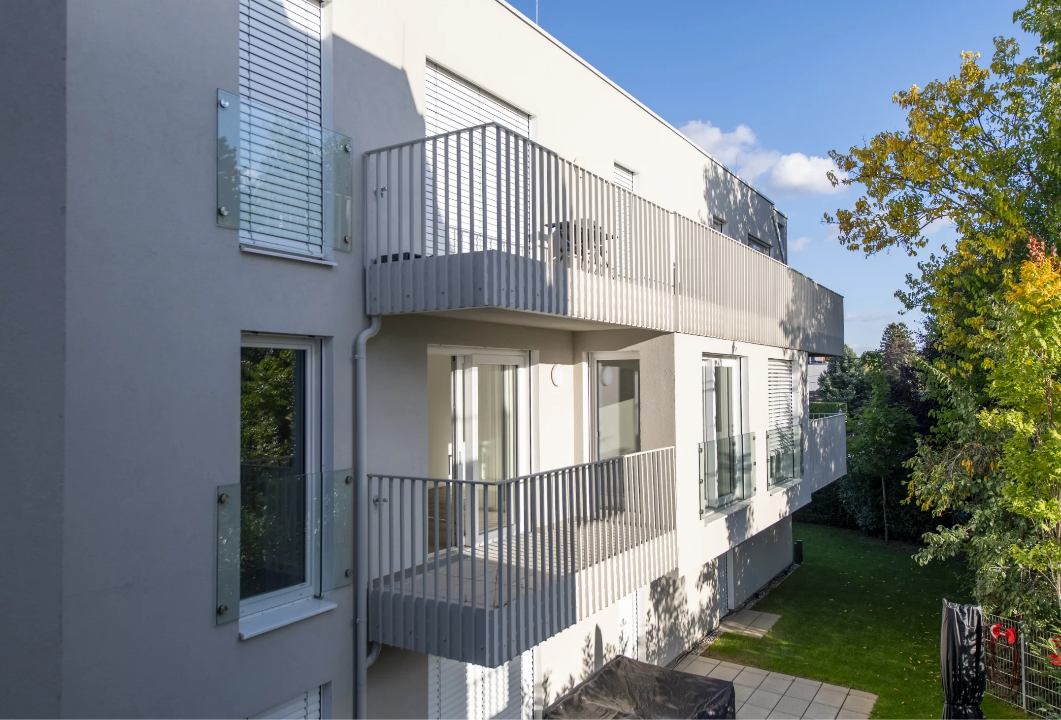 Die Außenansicht einer modernen, weißen Wohnanlage, die gestaffelte Balkone mit weißen Gittergeländern und eine angrenzende Grünfläche zeigt.